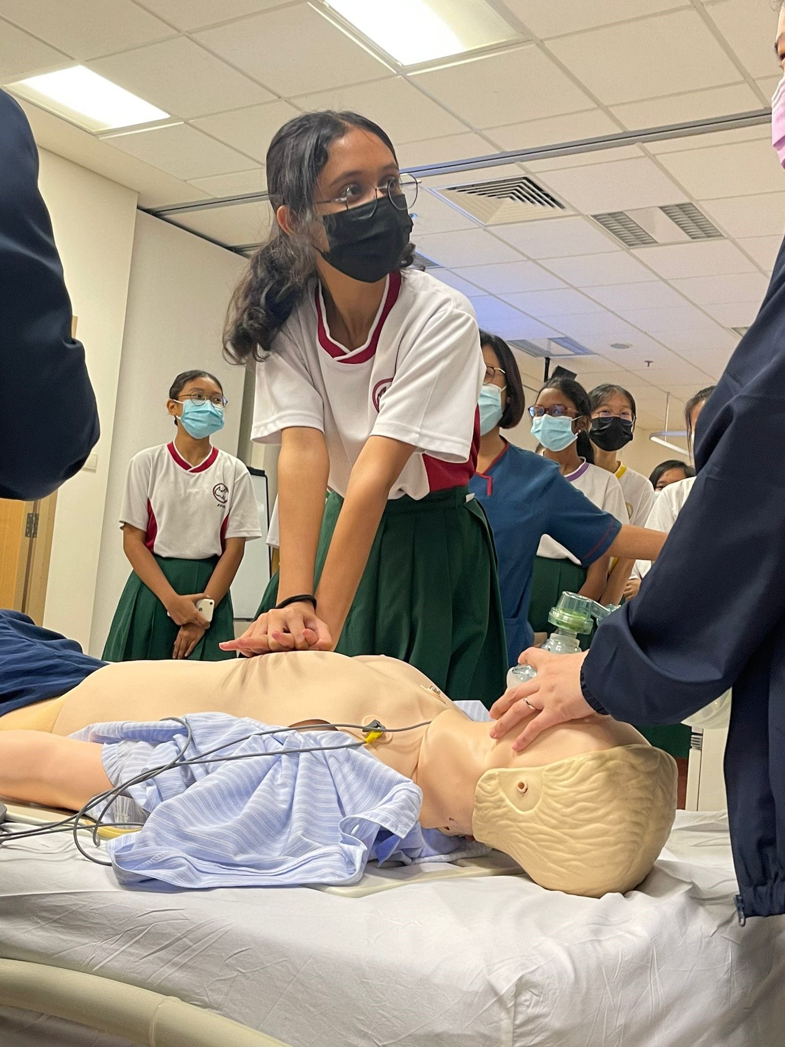 A masked schoolgirl in uniform performs chest compressions on a CPR mannequin while classmates watch indoors.